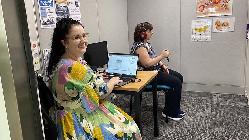 Woman (Audiologist) sitting at computer near another woman sitting with headphones on having their hearing assessed