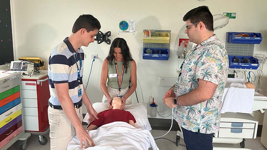 Male senior doctor standing watching one male and one female junior doctor doing training using a mannequin on a hospital bed