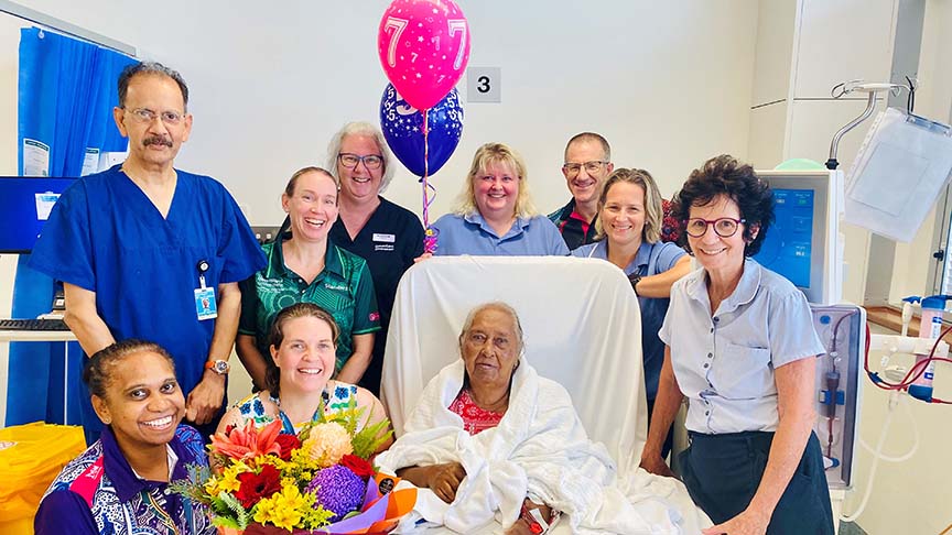 Woman (Elsie) sitting in a hospital bed surrounded by her care team who are holding balloons and flowers