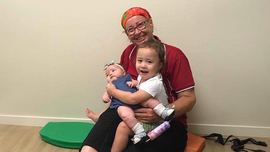 Little girl and baby sitting on older woman's lap on the floor smiling at camera